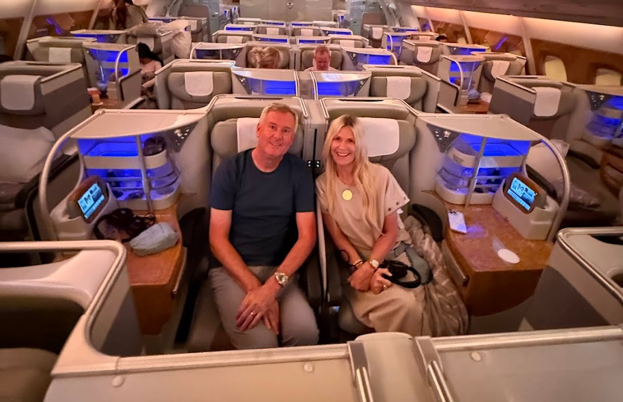 A Gen X couple seated side-by-side in a spacious Business Class cabin, smiling and relaxed, surrounded by sleek gray seats, woodgrain tables, and ambient blue lighting.