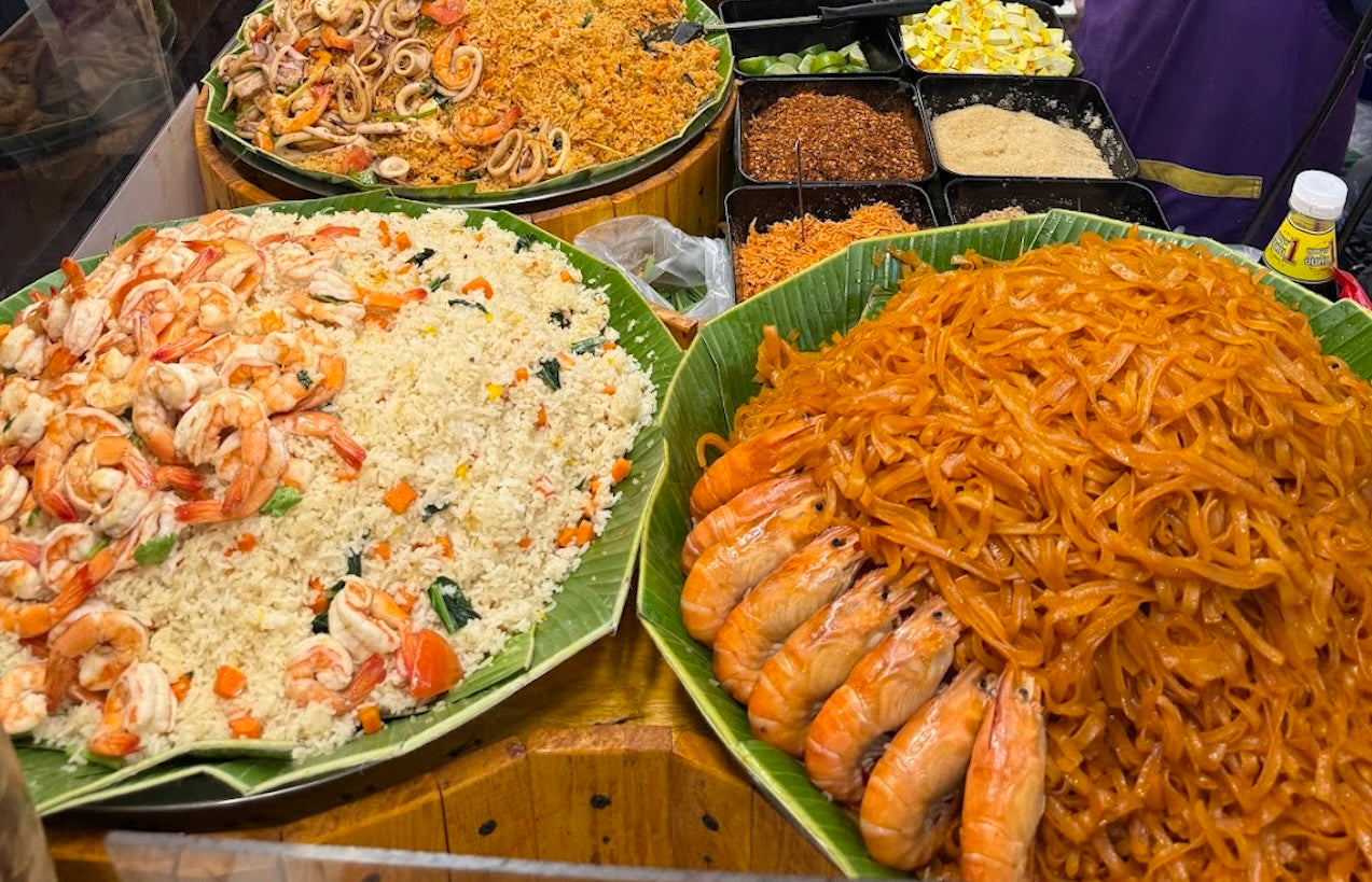 Thai street food display with mounds of Pad Thai, shrimp fried rice, and spicy seafood noodles in a Bangkok night market stall.