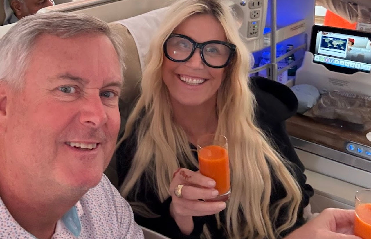 Couple smiling in Emirates Business Class cabin, holding glasses of orange juice with in-flight screens in the background.