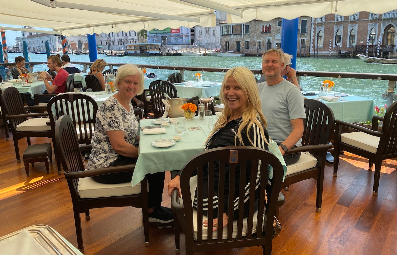 Three travelers seated at a canal-side restaurant in Venice, Italy, smiling at the table with the Grand Canal and historic buildings in the background.
