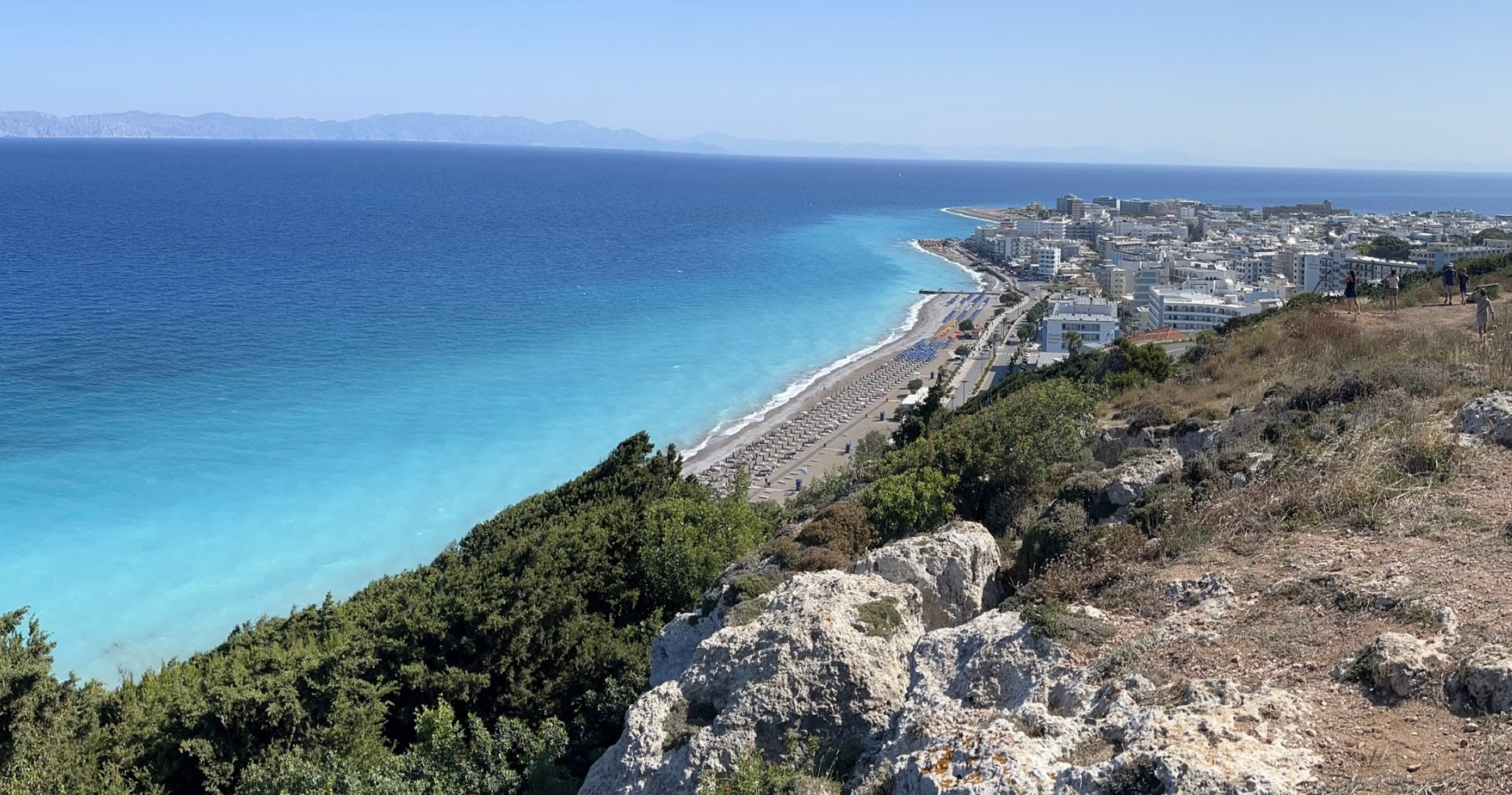 Panoramic view from Monte Smith in Rhodes, Greece, showing the turquoise Aegean Sea, coastline, and the city of Rhodes in the distance.
