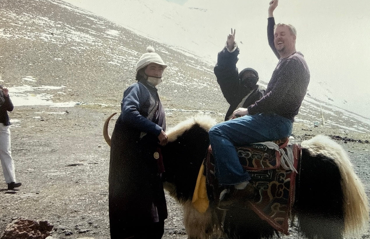 Traveler riding a decorated yak at high altitude in Tibet with a local guide, surrounded by rocky terrain and snow patches.