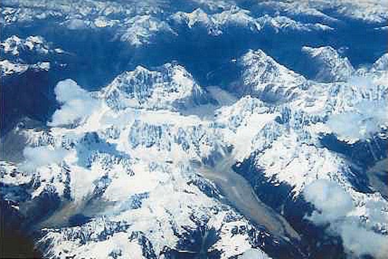 Aerial view of snow-covered Himalayan mountains with deep valleys and jagged peaks, seen from an airplane window en route to Tibet.
