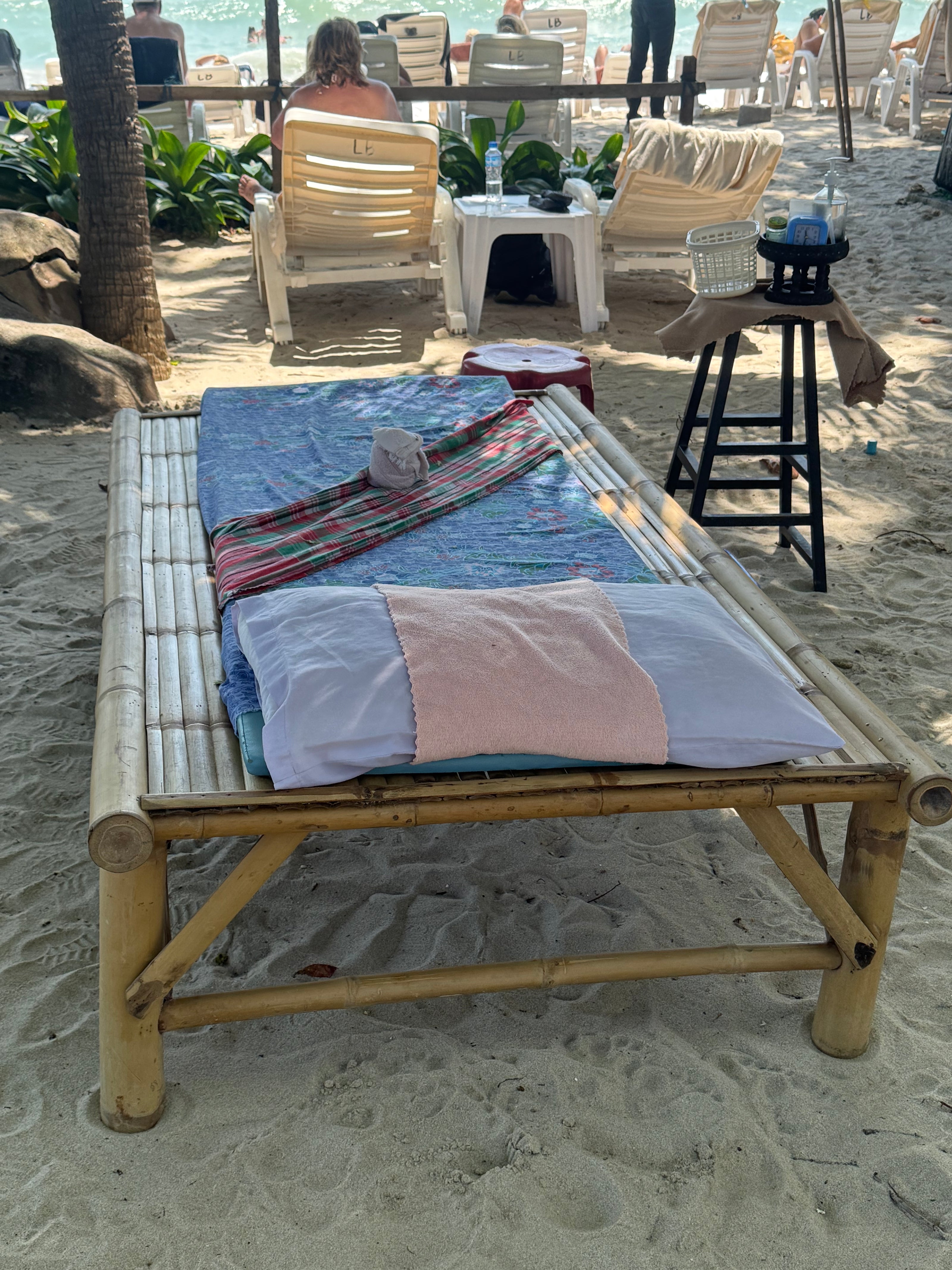 Simple bamboo massage bed on the beach, set up for a Thai massage under shade near the ocean, with beach chairs and sunbathers nearby.