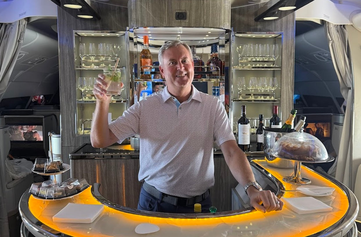 Man enjoying a cocktail at the Emirates A380 onboard bar, surrounded by glassware, bottles of wine and spirits, and ambient lighting in a premium cabin setting.