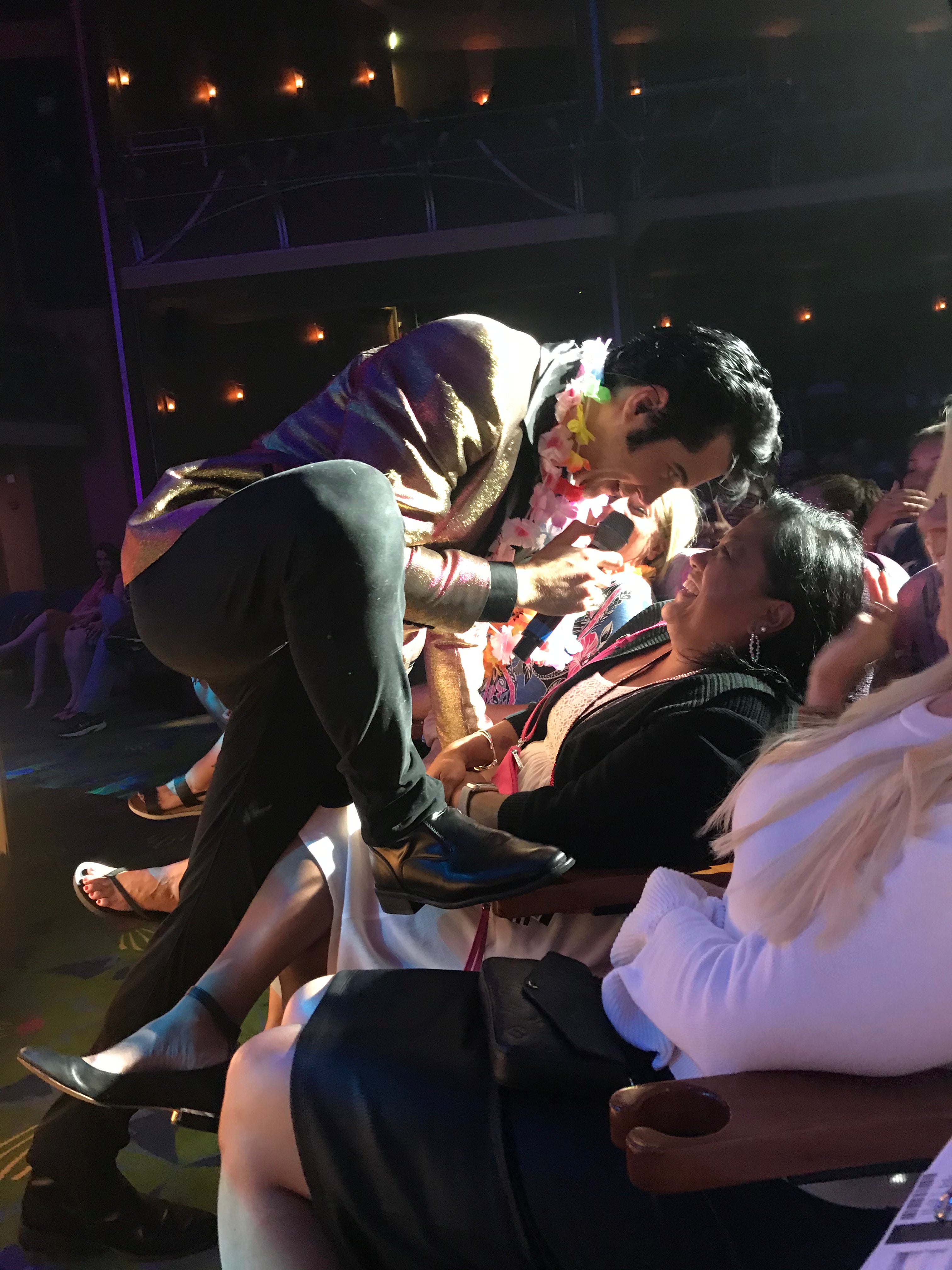 Performer in an Elvis costume on a cruise ship stage interacting with an audience member, with others in the crowd watching and smiling.