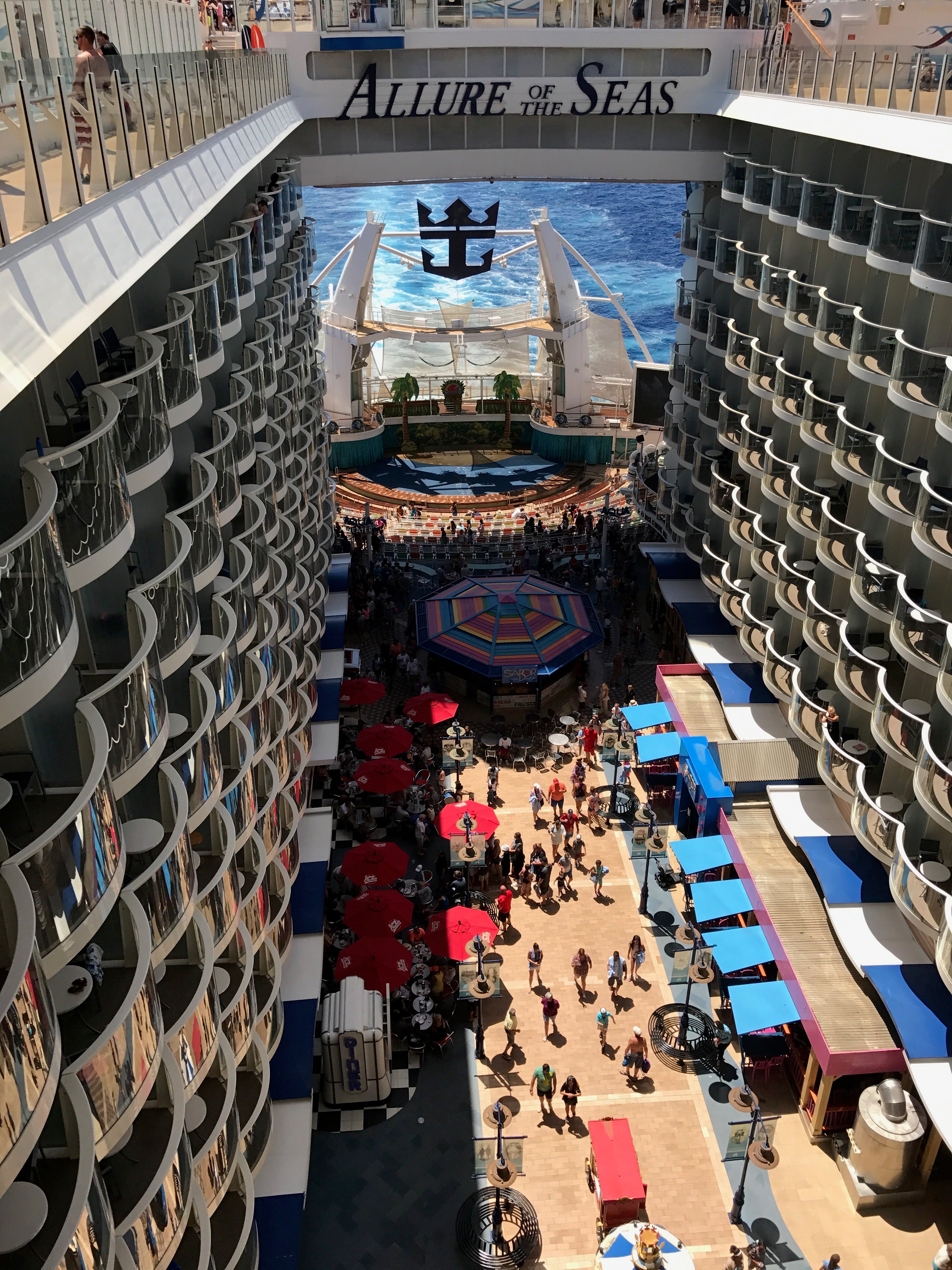 Overhead view of the Allure of the Seas' open-air Boardwalk neighborhood, featuring balconies, red umbrellas, shops, and the AquaTheater at the ship’s aft.