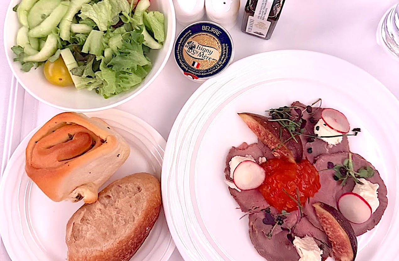 A gourmet in-flight meal served on white plates, featuring roast beef with figs, radish slices, whipped cheese, and red pepper relish, alongside a cucumber salad, two types of bread, and French butter.